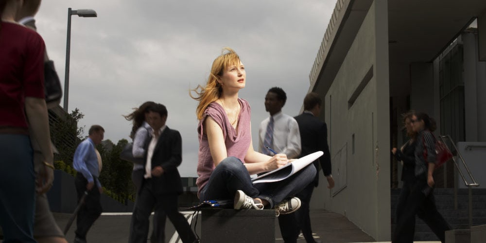 Young woman sitting cross legged in busy square with sketchpad